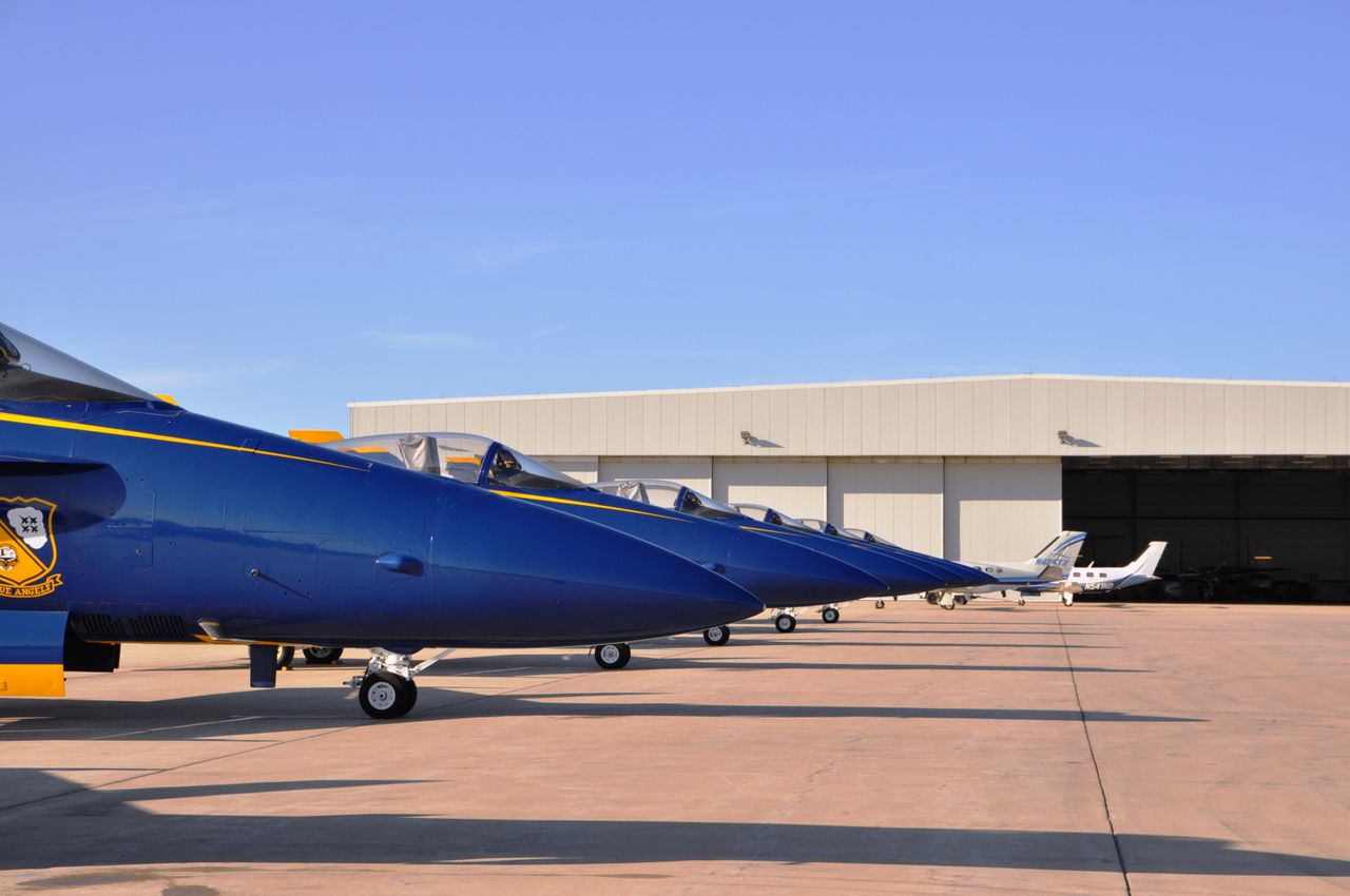 gallery-2 Blue fighter jets aligned on the tarmac in front of an aircraft hangar on a clear day.